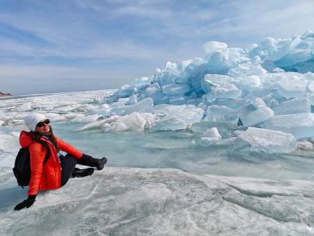 A woman enjoys the beauty of a large blue glacier while seated on the ice, surrounded by winter...