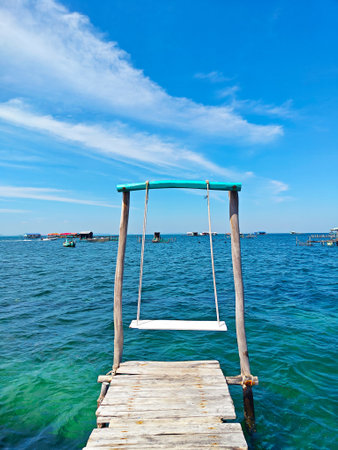 Wooden pier with a rustic swing extends over the turquoise sea under a bright blue sky.の写真素材