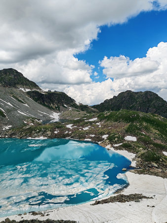 Alpine lake showcases vibrant turquoise water reflecting clouds and mountains, surrounded by snow patches and rocky terrainの写真素材