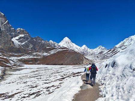 Trekking adventurers navigate snowy terrain surrounded by towering mountains and blue sky. Nepal,...