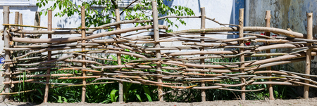 A braided wooden fence made of thin old branches in the countryside against the backdrop of an old houseの写真素材
