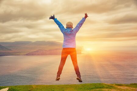 Young Woman Jumping At Cliff Edge, Sunrise Over Mountain, Kalsoy Island, Faroe Islandsの写真素材
