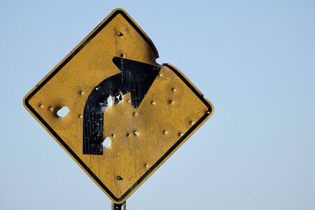 A street sign warning of a curve ahead is riddled with bullet holes.の写真素材