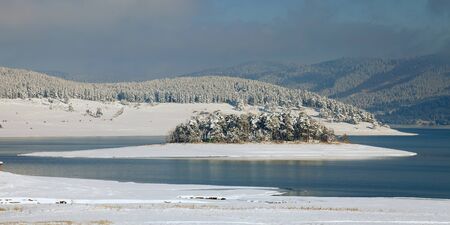 Unfrozen lake in the winter forests of Rhodopes, Bulgaria. Blue cold water and snow islandの写真素材