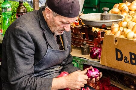 Old man sitting on a chair and he is peeling red onion in the Old Bazaar in Skopje のeditorial素材