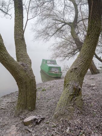 An old boat moored along the river bank in a willow grove. Dense fog in the river valley and frozen nature in a morning. A gloomy autumn landscape of frozen nature in a cold and silent fog.の写真素材