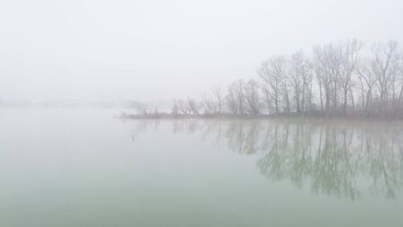 River island in dense fog, with tree silhouettes of bare branches in late fall. The usual autumn morning by the river. Mystical and melancholic autumn landscape.の写真素材