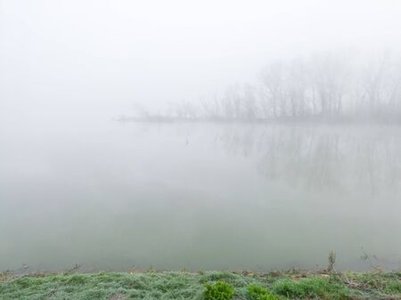 The gloomy landscape of the morning mist in the river valley. A thick fog hovers over the flat surface of the river and creeps among the trees in the woods on a barely visible river island.の写真素材