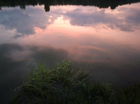 Abstract landscape with reflection of glowing sky and colorful clouds in water and green grass on...