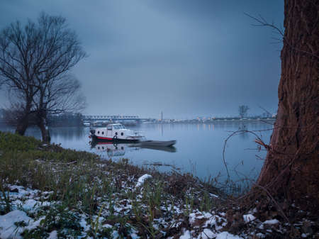 Moored boats near the shore during dusk in winter, bare trees and some snow on the bankの写真素材
