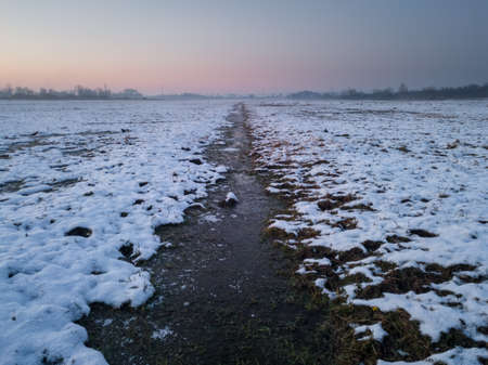 A shallow ditch with frozen water cuts across a meadow under snow at dusk, a mist in the distance and a purple glow in the skyの写真素材