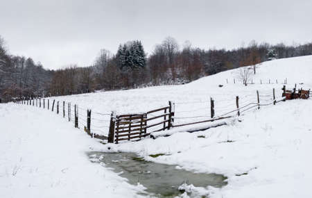 Hilly countryside in winter, empty animal pen against forestの写真素材