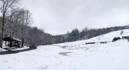 Hilly countryside in winter, empty animal pen against forest and stream, land covered with snowの写真素材