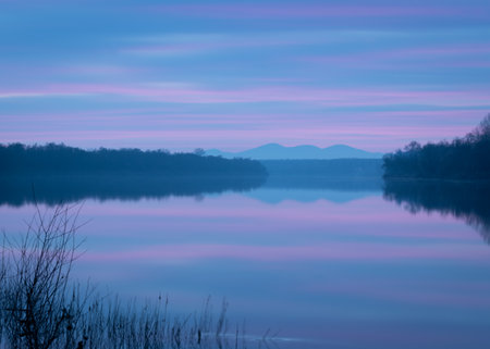 Peaceful atmospheric landscape with pastel colors, Sava river at twilight, forested banks lead to distant mountain silhouette in haze, calm nature and water reflectionの写真素材