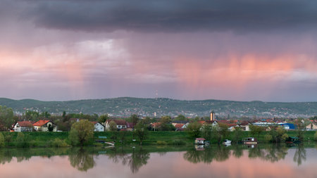 Strong downpour over hill behind settlement, sunlit clouds during sunsetの写真素材