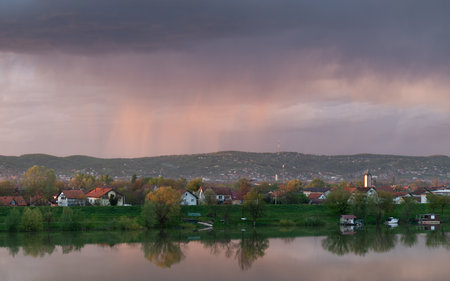Strong downpour over hill behind settlement, sunlit clouds during sunsetの写真素材