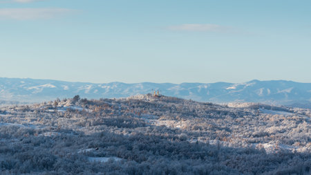 Hilly village covered with frost after cold winter night, there is church on top of hill surrounded with deciduous forest, mountain range in distanceの写真素材