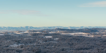 Hilly village covered with frost after cold winter night, rural landscape with houses, deciduous forest and fields, mountain range in distanceの写真素材