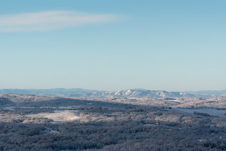 Hilly village covered with frost after cold winter night, rural landscape with houses, deciduous forest and fields, mountain range in distanceの写真素材