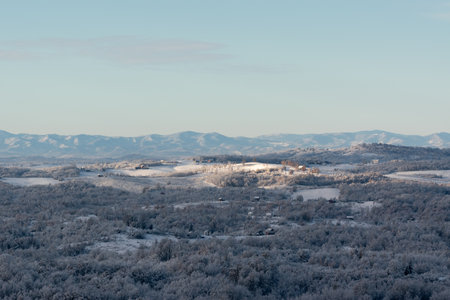 Hilly village covered with frost after cold winter night, rural landscape with houses, deciduous forest and fields, mountain range in distanceの写真素材
