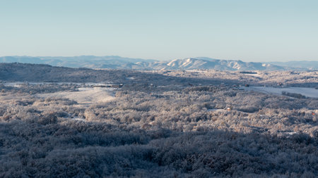 Hilly village covered with frost after cold winter night, rural landscape with houses, deciduous forest and fields, mountain range in distanceの写真素材