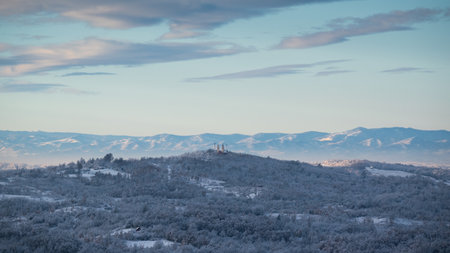Hilly village covered with frost after cold winter night, there is church on top of hill surrounded with deciduous forest, mountain range in distanceの写真素材