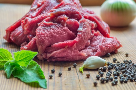 Piece of beef meat in preparation on wooden desk with black pepper, onion, garlic and mint leaves.の写真素材