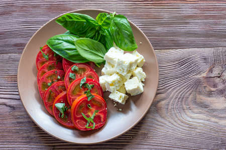 Ripe tomatoes, feta cheese and basil leaves in clay plate on dark wooden background.の写真素材