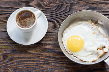 Fried egg on the pan and white  cup of coffee for breakfast. Wooden table, Top view.の写真素材