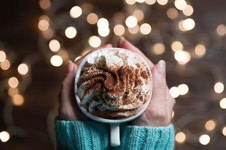 Woman hands holding white ceramic mug of hot chocolate with whipped cream in hands. Blurred Christmas lights at background, copy space.の写真素材