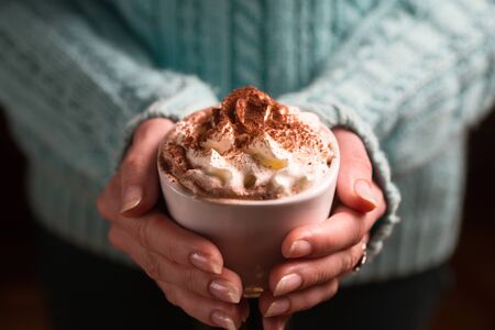 Female hands holding white ceramic mug of hot chocolate with whipped cream in hands wearing  green...
