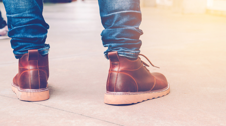 Young men fashion in leather boots walk in urban city. Close up view on man's legs in blue jeans and brown leather boots with sunlight against tower building blur background for copy space. の写真素材