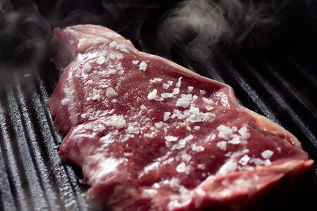 A slice of sirloin steak frying in a griddle pan. The steak is seasoned with salt and smoke is rising from the heat of the pan.の写真素材