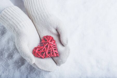 Female hands in white knitted mittens with a entwined vintage romantic red heart on a winter snow...