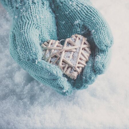Woman hands in light teal knitted mittens are holding a beautiful entwined vintage romantic red heart in a snow background.の写真素材