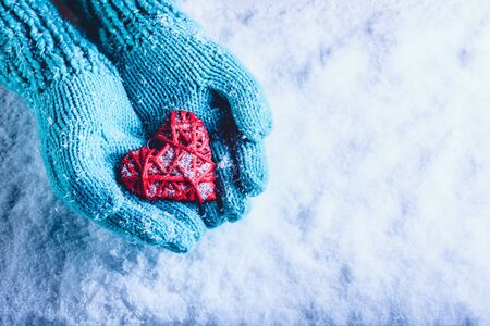 Woman hands in light teal knitted mittens are holding a beautiful entwined vintage romantic red heart in a snow background.の写真素材