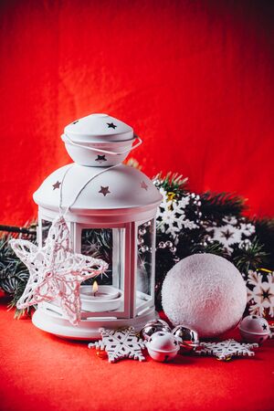 Magical white lantern is standing with beautiful white knit star on it and a fir tree branch and a snowball lying in front of it on a christmas red background. Christmas card with copy space.の写真素材
