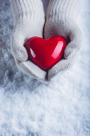 Female hands in white knitted mittens with a glossy red heart on snowの写真素材