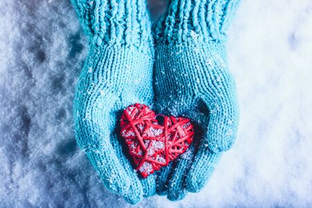 Woman hands in light teal knitted mittens are holding a beautiful entwined vintage romantic red heart in a snow background.の写真素材