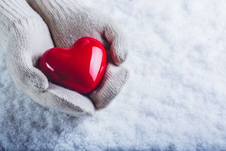 Female hands in white knitted mittens with a glossy red heart on snowの写真素材