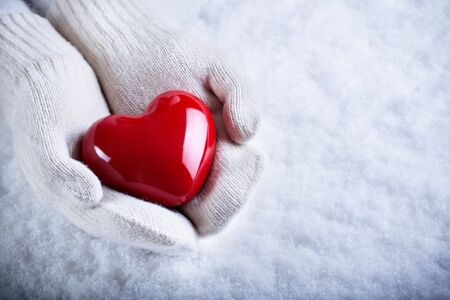 Female hands in white knitted mittens with a glossy red heart on a snow background.の写真素材