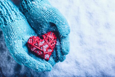 Woman hands in light teal knitted mittens are holding a beautiful entwined vintage romantic red heart in a snow background.の写真素材