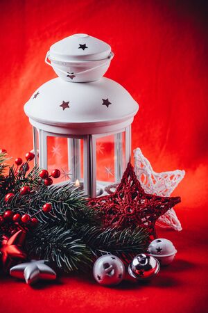 Christmas white lantern is standing with a burning candle in it with a fir tree branch and knit stars in front of it on a red background.の写真素材