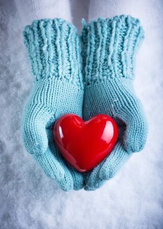 Female hands in light teal knitted mittens with a glossy red heart on snowの写真素材