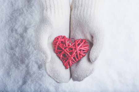 Female hands in white knitted mittens with a entwined vintage romantic red heart on a winter snow background.の写真素材