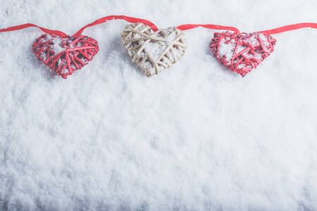 Three beautiful romantic vintage hearts are hanging on a red band on a white snow winter background.の写真素材