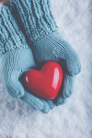 Female hands in light teal knitted mittens with a glossy red heart on snowの写真素材