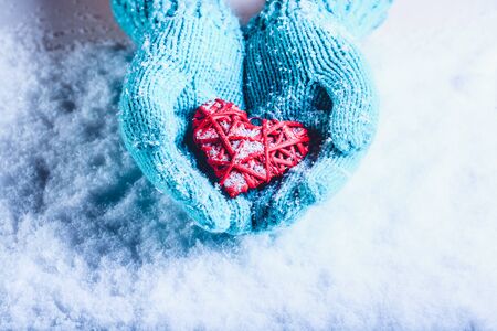 Woman hands in light teal knitted mittens are holding a beautiful entwined vintage romantic red heart in a snow background.の写真素材