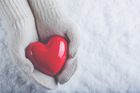 Female hands in white knitted mittens with a glossy red heart on a snow winter background.の写真素材