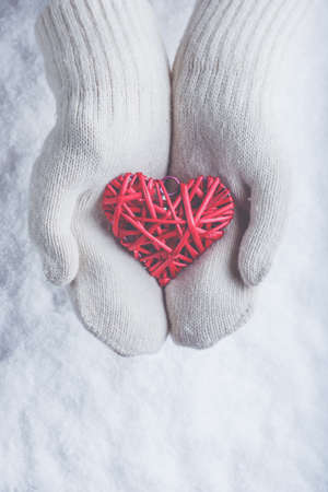 Female hands in white knitted mittens with a entwined vintage romantic red heart on a winter snow background.の写真素材